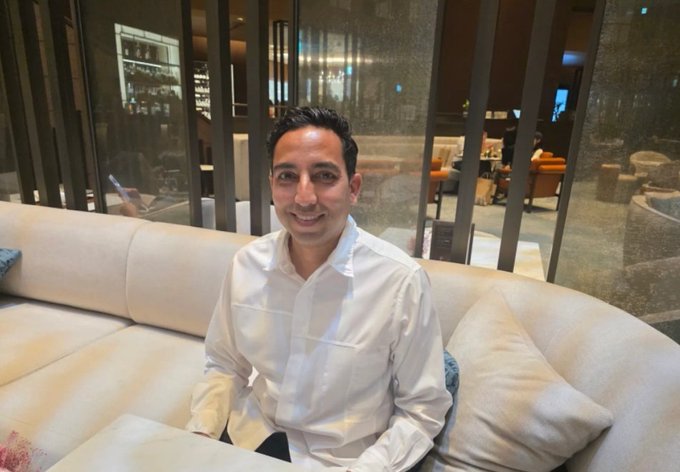 A man in a white dress shirt smiles while seated on a beige couch in a modern lounge setting, pictured during an interview with SBI-backed Ripple-affiliated Evernorth, which is establishing a Korean unit and pursuing a KRX listing for XRPN.