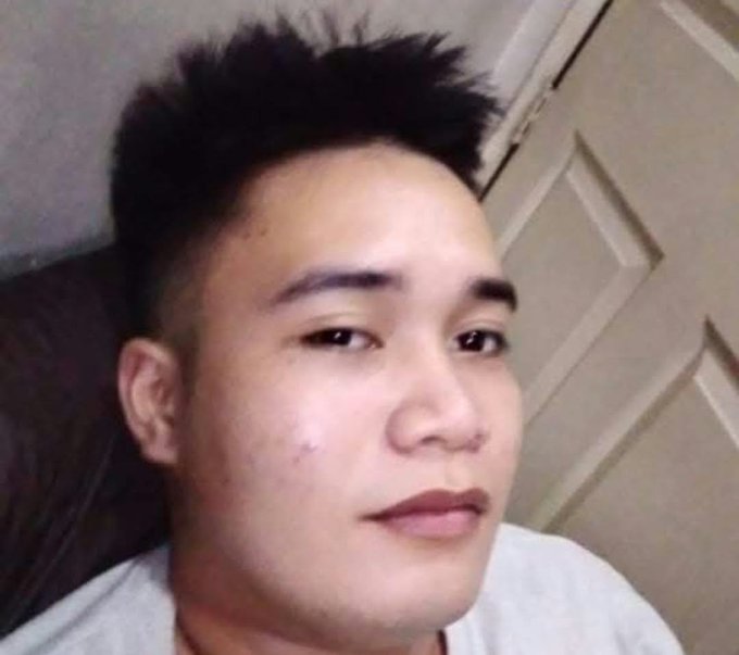 A close-up selfie of a young Asian man with short, spiky black hair wearing a white shirt, positioned against a light-colored paneled door.