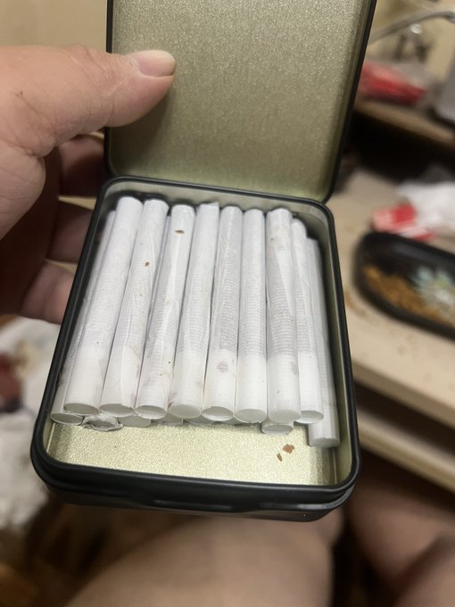 A hand holds an open tin box containing several hand-rolled white cigarettes.