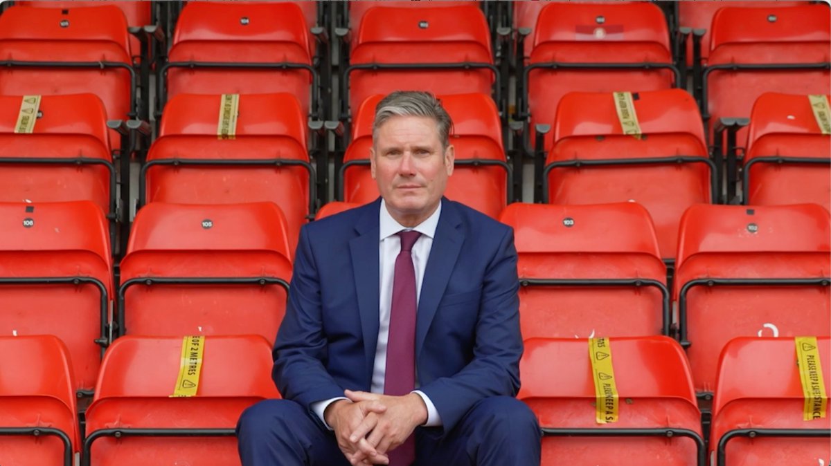 A man in a navy blue suit and maroon tie sits in front of rows of empty red stadium seats marked with yellow social distancing tape reading 'DO NOT SIT HERE' and 'KEEP YOUR DISTANCE'.