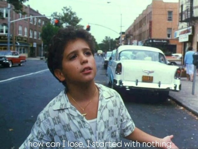 A young boy in a patterned shirt stands on a city street with vintage cars behind him; subtitle reads 'how can I lose, I started with nothing'.