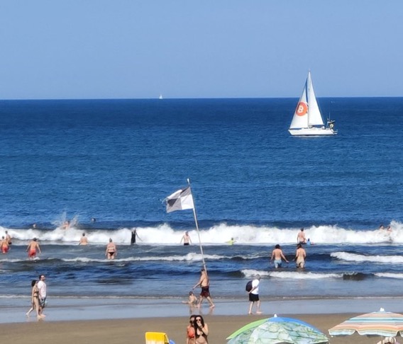 A sunny beach scene shows people swimming and walking along the shore while a white sailboat glides across the blue ocean.