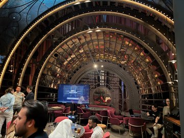 Interior view of a lavishly decorated venue with arched, illuminated ceiling structures and a stage with screens, where people are seated in a lounge-style setting with small tables.