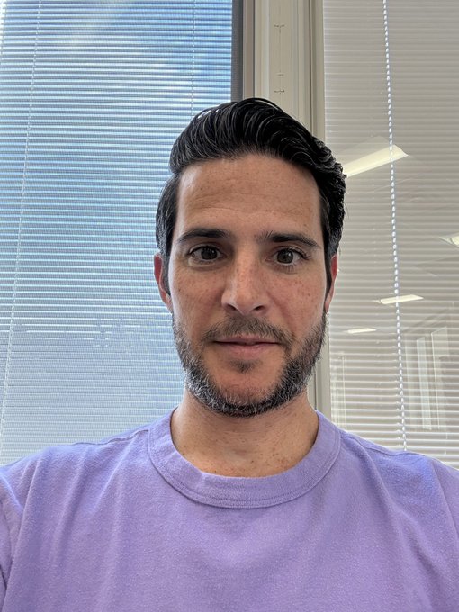 A man with dark hair and a salt-and-pepper beard wearing a light purple t-shirt poses for a selfie in front of window blinds, looking directly at the camera with a neutral expression—appearing to be a casual profile photo accompanying a post seeking to connect with fellow crypto/NFT enthusiasts who collect digital art.