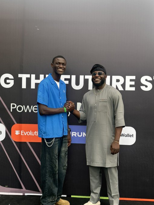 Two men posing together at an event with a backdrop reading '...G THE FUTURE S...', 'Power...' and 'Evolutio... Wallet', one in a blue shirt and the other in a gray traditional outfit with sunglasses.