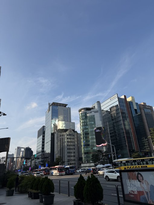 A street view of a city with modern skyscrapers and digital billboards under a partly cloudy sky, with a 'CHAGEE' ad visible.