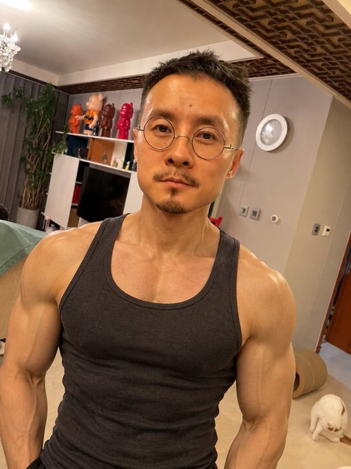 A muscular man in a gray tank top poses in his home, showing off his toned arms and chest. The image reflects the post's focus on a fit physique, with the caption humorously referencing the "slim and toned look" trend and playfully noting that this appearance wouldn't suit blond-haired individuals.