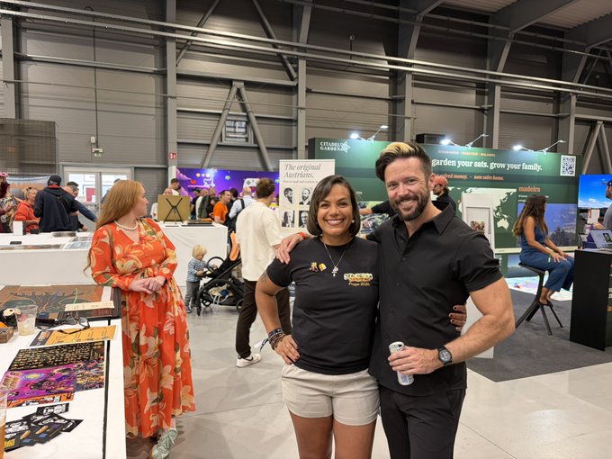 The image shows a man and woman standing together, posing for a photo at an indoor event. They are both smiling and appear to be enjoying themselves. The background of the image reveals that they are surrounded by other people who seem to be engaged in conversations or activities as well. There is also a TV visible in the scene, which might be displaying information related to Bitcoin or the event itself.