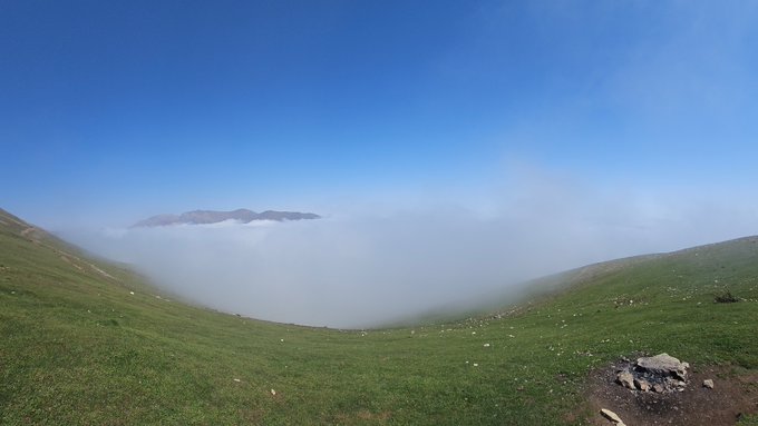 A misty mountain landscape with green rolling hills in the foreground and a thick layer of fog filling the valley below a clear blue sky.