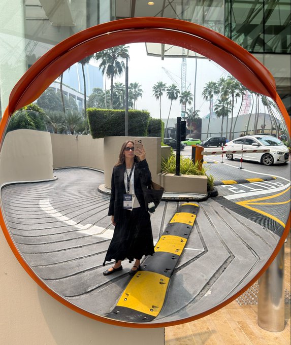 A fisheye mirror selfie of a woman in a black outfit with a lanyard, standing at a parking garage entrance with palm trees visible in the background.