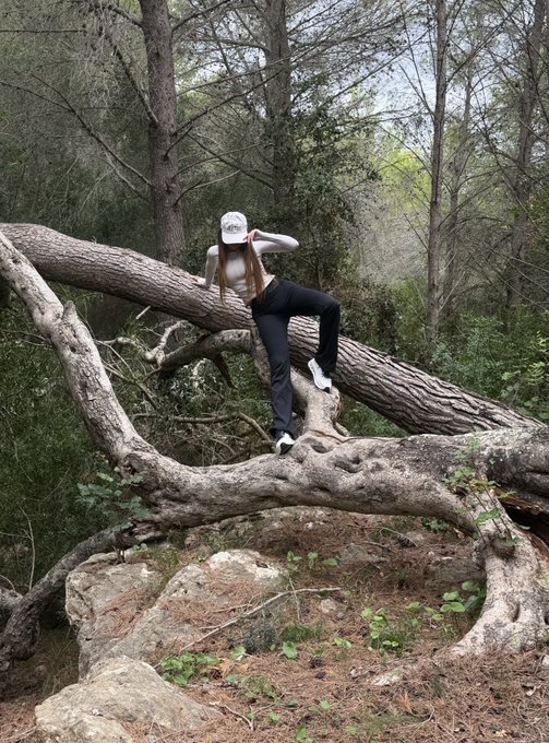 A person wearing a white cap, white long-sleeve top, and black pants poses on a large fallen tree in a wooded outdoor area.