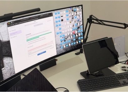 A home office setup featuring a large curved monitor displaying what appears to be order management software, alongside a tablet on a stand and keyboard.
