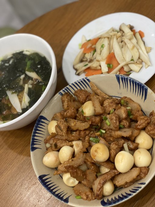 A homemade Vietnamese meal spread of stir-fried pork with quail eggs in a blue-rimmed bowl, alongside a side of sautéed mushrooms with carrots and a bowl of seaweed soup, perfectly matching the cozy "CƠM NHÀ" (home-cooked meal) theme inviting family to share the meal despite the rainy weather.