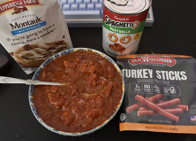 A bowl of chili or soup sits next to a can of Campbell's SpaghettiOs Meatballs, a bag of Pepperidge Farm Montauk milk chocolate cookies, and a package of Old Wisconsin Turkey Sticks smoked sausage.