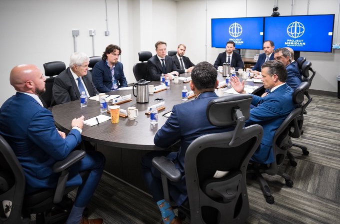 A group of businessmen in suits sit around a conference table in a meeting room, with two screens in the background displaying 'PROJECT MERIDIAN'.
