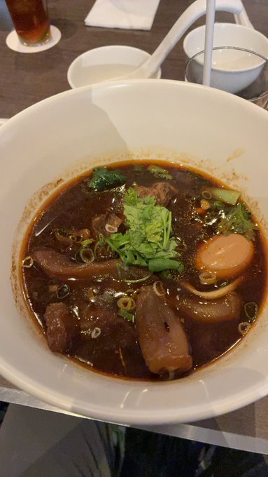 A bowl of dark broth noodle soup topped with greens, meat, and a halved boiled egg is served on a table.