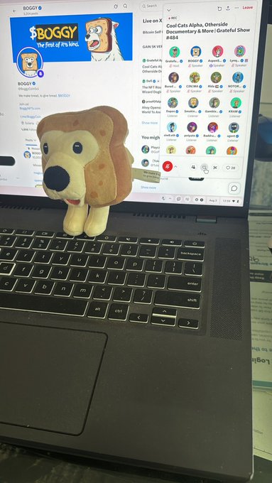 A laptop displaying the Twitter/X interface showing the 'BOGGY' cryptocurrency profile and a Live on X feed for an NFT show, with a cute brown dog plush toy sitting on the keyboard.
