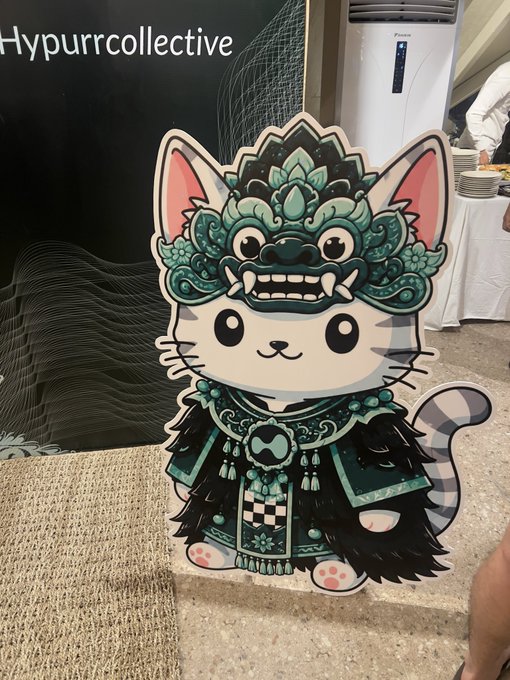 A cartoon cutout of a cute white cat wearing an elaborate teal and black samurai-style armor with a fierce faceplate, displayed at what appears to be a convention or store, with "HypurrCollective" partially visible in the upper left.