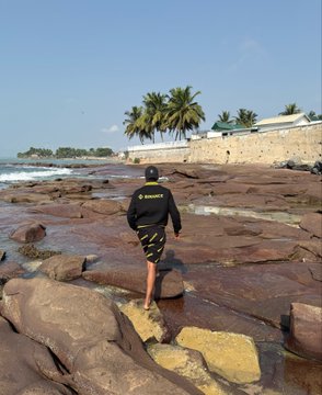 The image shows a person walking across rocks, possibly on a beach or near a body of water. The person appears to be wearing shorts and carrying a backpack while navigating through the terrain. The surrounding area features palm trees, indicating that they are likely in a tropical location. The caption "One step at a time. Just don't stop! #KeepBuilding @cz_binance @binance @BinanceAfrica @BinanceAngels" suggests that the person is on an adventure or journey and should continue to move forward despite any obstacles or challenges they may face along the way.