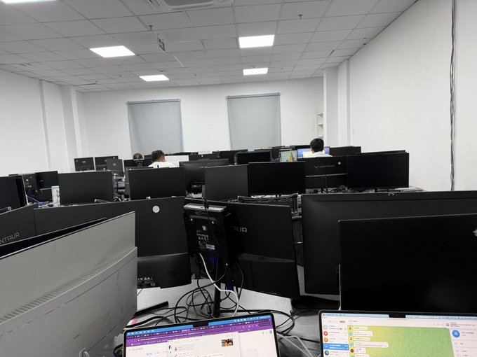 The image shows a large office space with multiple computer monitors, laptops, and people working at their desks. There are several computers placed throughout the room, including desktop computers and laptop computers. The workers appear to be focused on their tasks, possibly engaged in various projects or collaborating on different assignments. The overall atmosphere suggests that this is an active workspace where individuals come together to complete work-related tasks.
