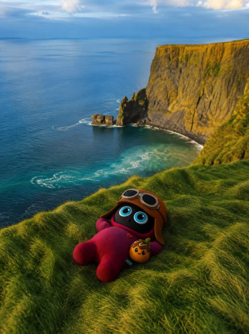 A small plush toy wearing an aviator cap and goggles lies on grassy cliffs overlooking the ocean and steep cliffs.