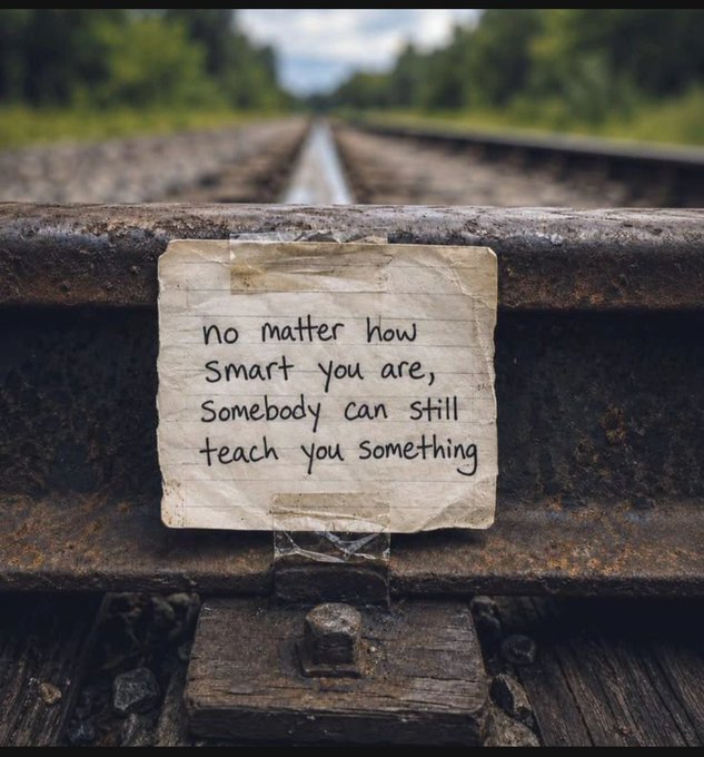 A handwritten note taped to a rusted railroad track reads 'no matter how smart you are, somebody can still teach you something', with the blurred tracks and trees receding into the distance.
