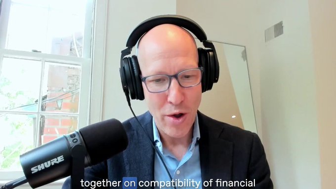 A bald man with glasses wearing headphones and a dark blazer speaks into a Shure microphone, with the subtitle 'together on compatibility of financial' visible at the bottom.