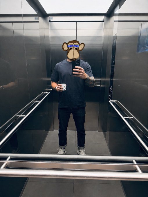 A person takes a mirror selfie in an elevator, wearing a Bored Ape virtual mask/filter, dressed in a casual grey t-shirt, dark jeans, and white sneakers while holding a smartphone. The image visually embodies the post's statement, with the Bored Ape filter symbolizing membership in the Bored Ape Yacht Club and the proud declaration of being part of the best community on the internet.