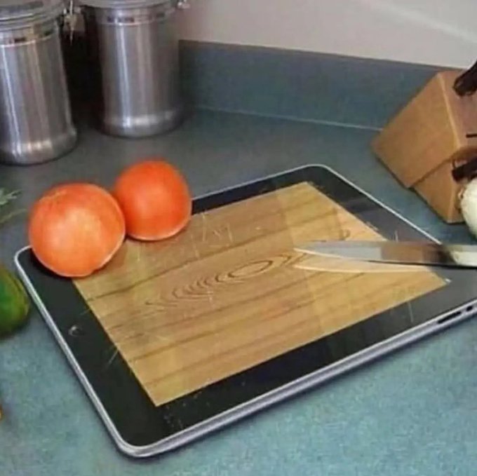 Two whole tomatoes, a lime, and a knife on a wooden cutting board placed on top of a laptop, alongside a knife block and metal canisters on a kitchen counter.