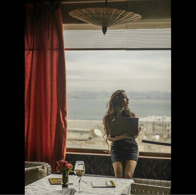 A woman with long wavy hair stands by a window overlooking a cityscape and water, holding a folder; a table with a wine glass and flowers is in the foreground.