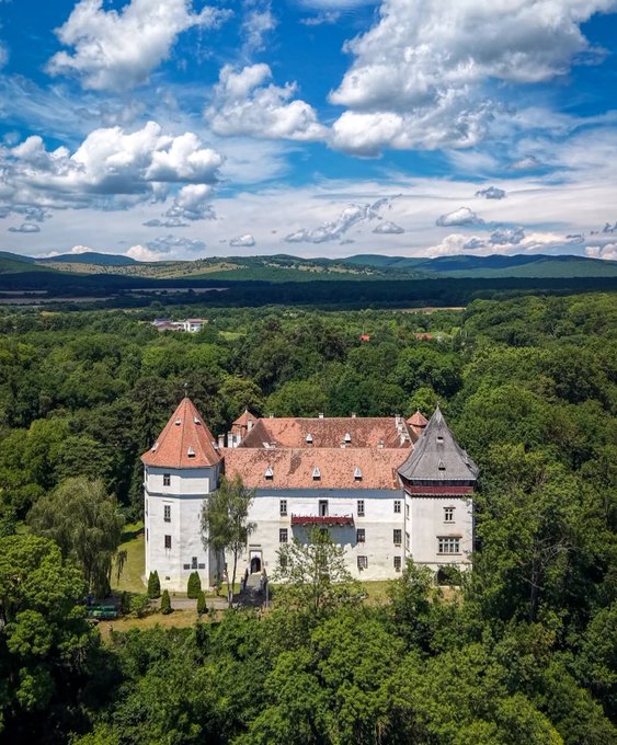 An aerial photograph of a large white castle or manor with a red tile roof and towers, situated in a lush green forested landscape with rolling hills and a partly cloudy sky in the background.
