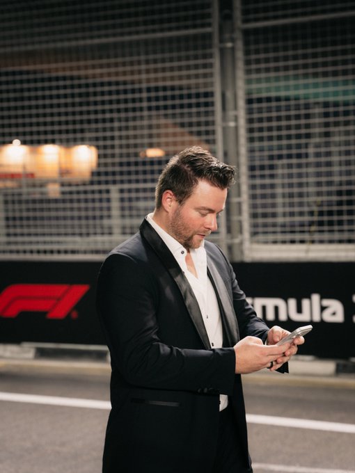 A man in a black tuxedo stands on the Formula 1 track in Singapore, looking down at his phone with the iconic "F1" and "Formula" branding visible on the barrier behind him.