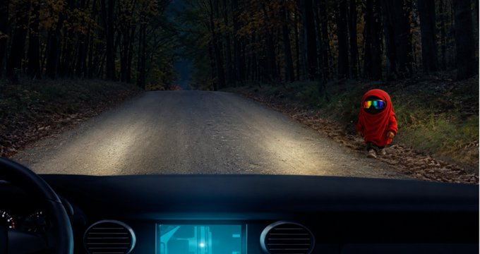 A view from inside a car driving down a dark forest road at night, with headlights illuminating the path and a figure in a red hooded jacket with a reflective visor standing on the right side of the road.