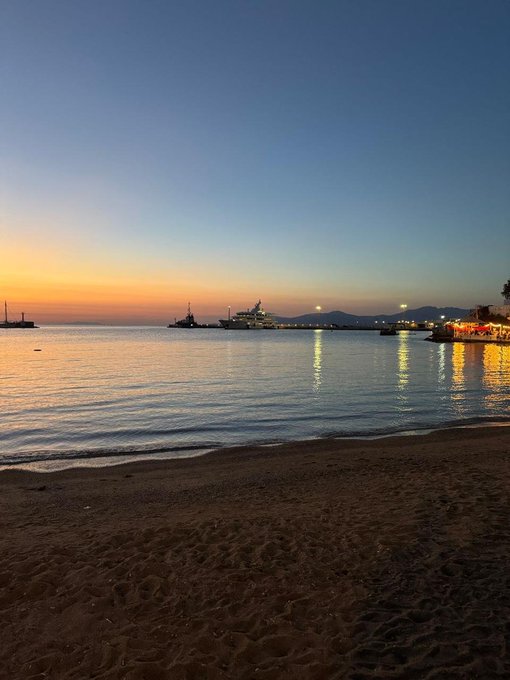 A serene beach scene at sunset or sunrise with calm water, a sandy shore in the foreground, boats and ships anchored in the harbor, and distant mountains under an orange and blue sky.
