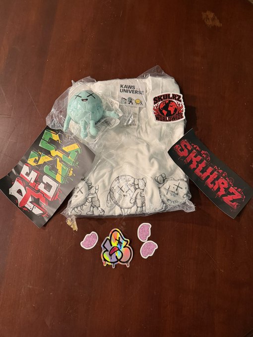 A collection of KAWS x Cactus Plant Flea Market merchandise including a t-shirt, plush toy, stickers, and cards with 'KAWS UNIVERSE' and 'SKULLRZ WORLDWIDE' branding, laid out on a wooden surface.