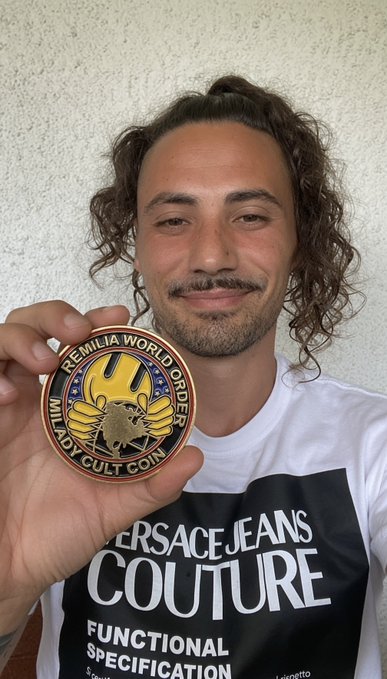 A man wearing a "VERSACE JEANS COUTURE FUNCTIONAL SPECIFICATION" shirt holds up a circular "MILADY WORLD ORDER CULT COIN" featuring a yellow cartoon character, proudly displaying his membership in the Milady Cult coin community.