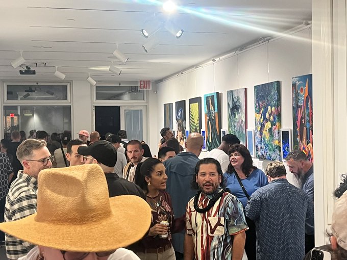 A crowded art gallery opening features attendees mingling in front of a wall displaying colorful paintings, including one signed 'FINTAN'