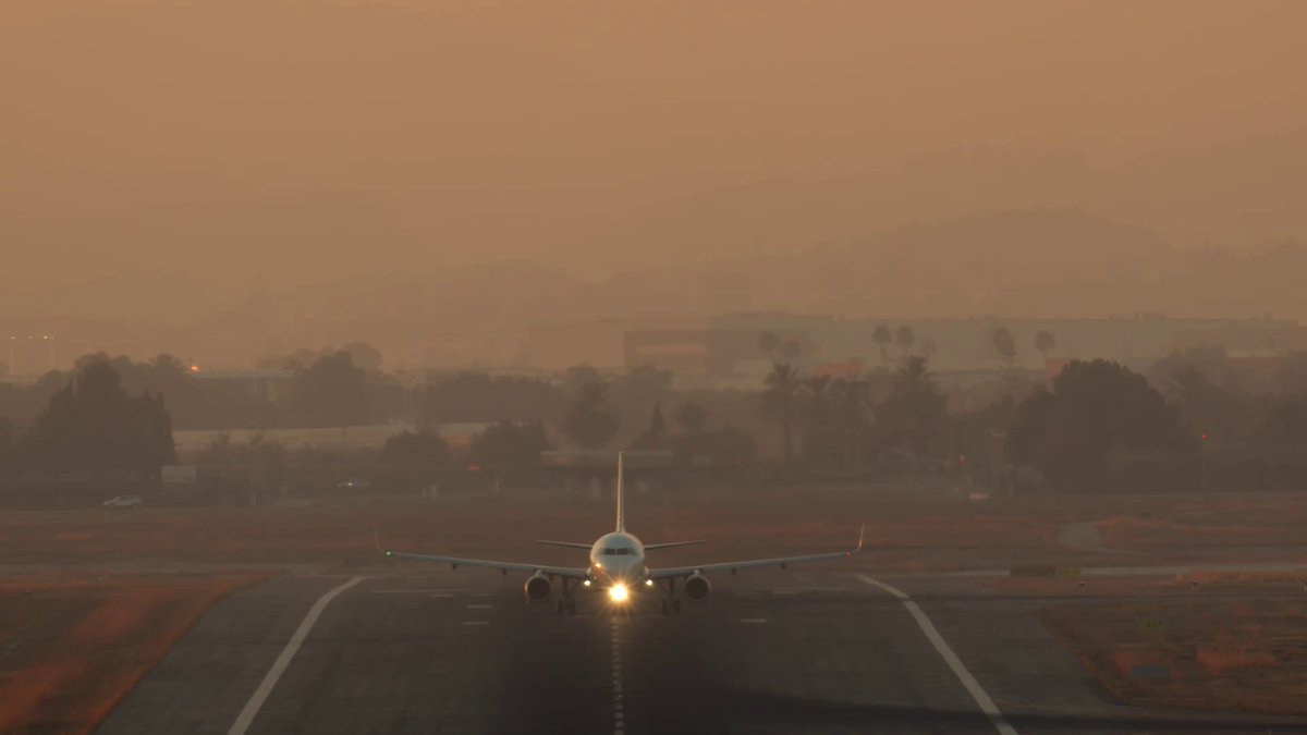 The image shows a large airplane taking off from an airport runway, with its landing gear down. The sky above appears hazy, suggesting that the photo was taken during sunset or early morning hours when visibility is lower due to atmospheric conditions.