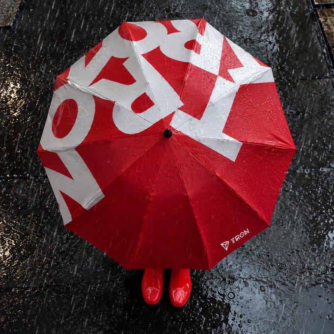 A top-down view of a person holding a red and white umbrella branded with the TRON logo while wearing red shoes on a wet pavement.