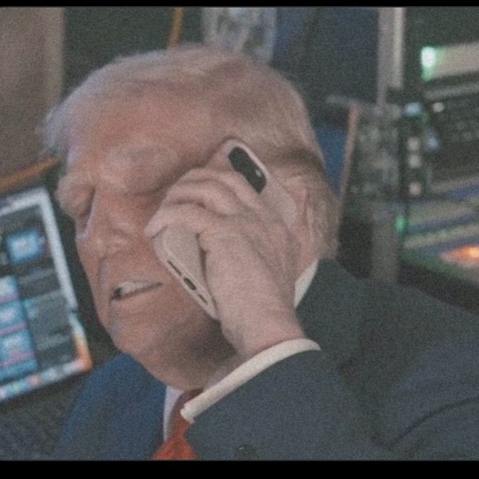 A man with light hair holds a smartphone to his ear with a concerned expression, with computer monitors and electronic equipment visible in the background.