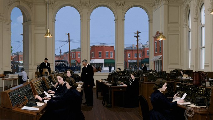 A vintage colorized photograph showing women operating telephone switchboards in a large room with tall arched windows. Through the windows, a city street with buildings and utility poles is visible.