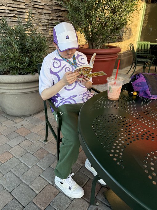 A young man wearing a white and purple cap, a white t-shirt with purple swirl patterns, and green pants, sitting at an outdoor patio table reading a Magic: The Gathering card while a pink drink sits nearby.