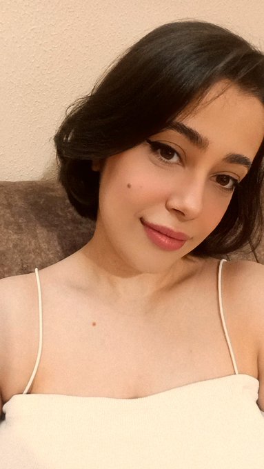 A young woman with short black hair and a white spaghetti-strap top poses for a close-up portrait, wearing subtle makeup with a small beauty mark on her cheek.