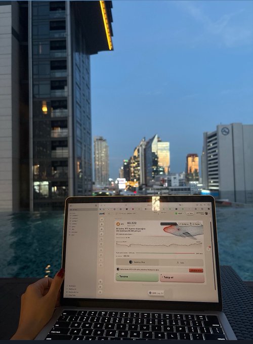 A person uses a laptop beside an outdoor pool overlooking a city skyline at dusk, with the stock chart “AAVE” showing a price around $83.322 on screen. The pleasant poolside setting matches the post’s “the weather is nice outdoors, $AAVE is going” message while the rising chart reinforces the bullish sentiment.
