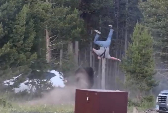 A person mid-flip in the air outdoors near trees, with a red metal box and a vehicle visible in the background, and dust rising from the ground.
