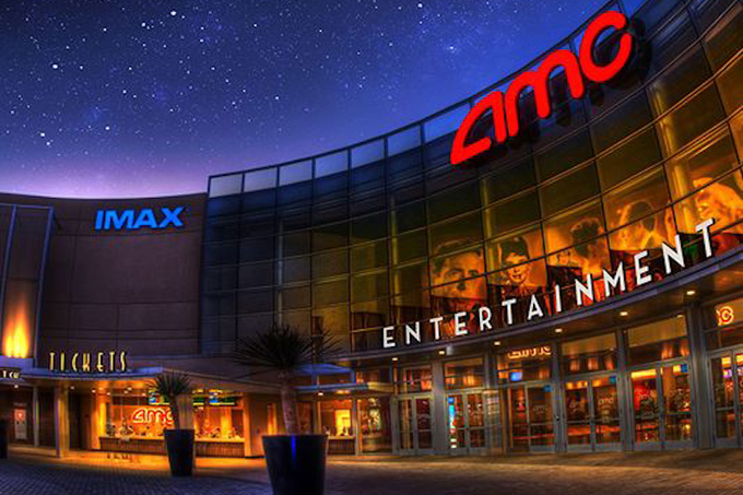 An AMC movie theater exterior is illuminated at dusk, displaying large red "AMC" signage, an "IMAX" sign, and the word "ENTERTAINMENT" across its glass facade, tying into AMC's record-breaking weekend of 10.2 million moviegoers and the company's new tokenized $AMC trading on Solana.
