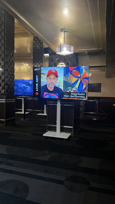 A digital display screen in a venue shows "LIVE ART" featuring "Senior Aquino" scheduled for "10am - 2pm Thursday," alongside another screen and an aquarium.