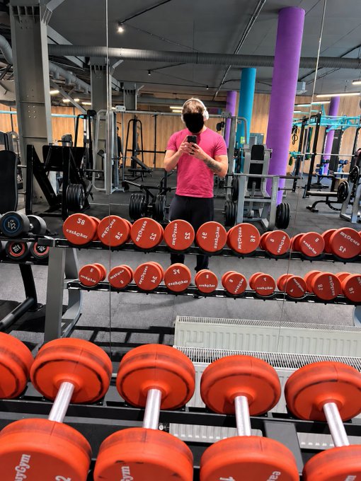 A man in a pink shirt takes a mirror selfie in a gym, surrounded by rows of orange Reebok dumbbells.