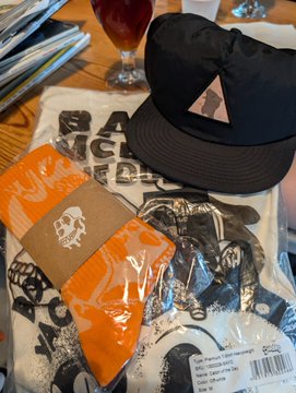 A flat lay photograph showing a black cap with a triangular logo, an orange packaged item with a skull design, placed on top of an enamel pin collection and printed materials on a wooden table.