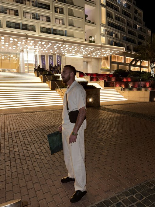 A man in white stands outside a grand, illuminated hotel at night, holding a shopping bag near the "FINAL" and "PMLA" banners, looking like he's ready to drop the best one-liner of the night to claim that 1 SOL prize.